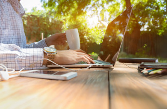 Man sitting at natural country style wooden desk with electronic gadgets around working on laptop drinking coffee sunlight and green terrace on background